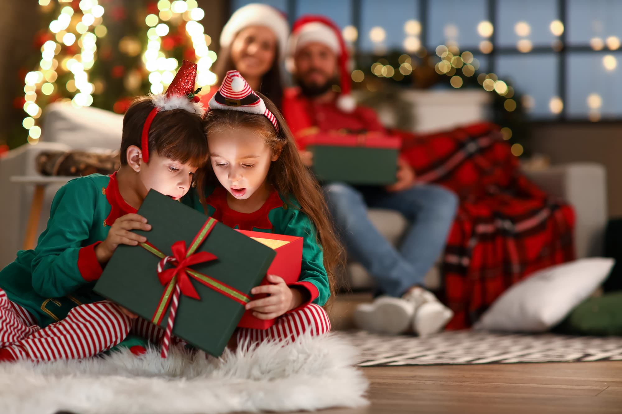 Two children open a present together at Christmas. Their parents look on smiling behind them.