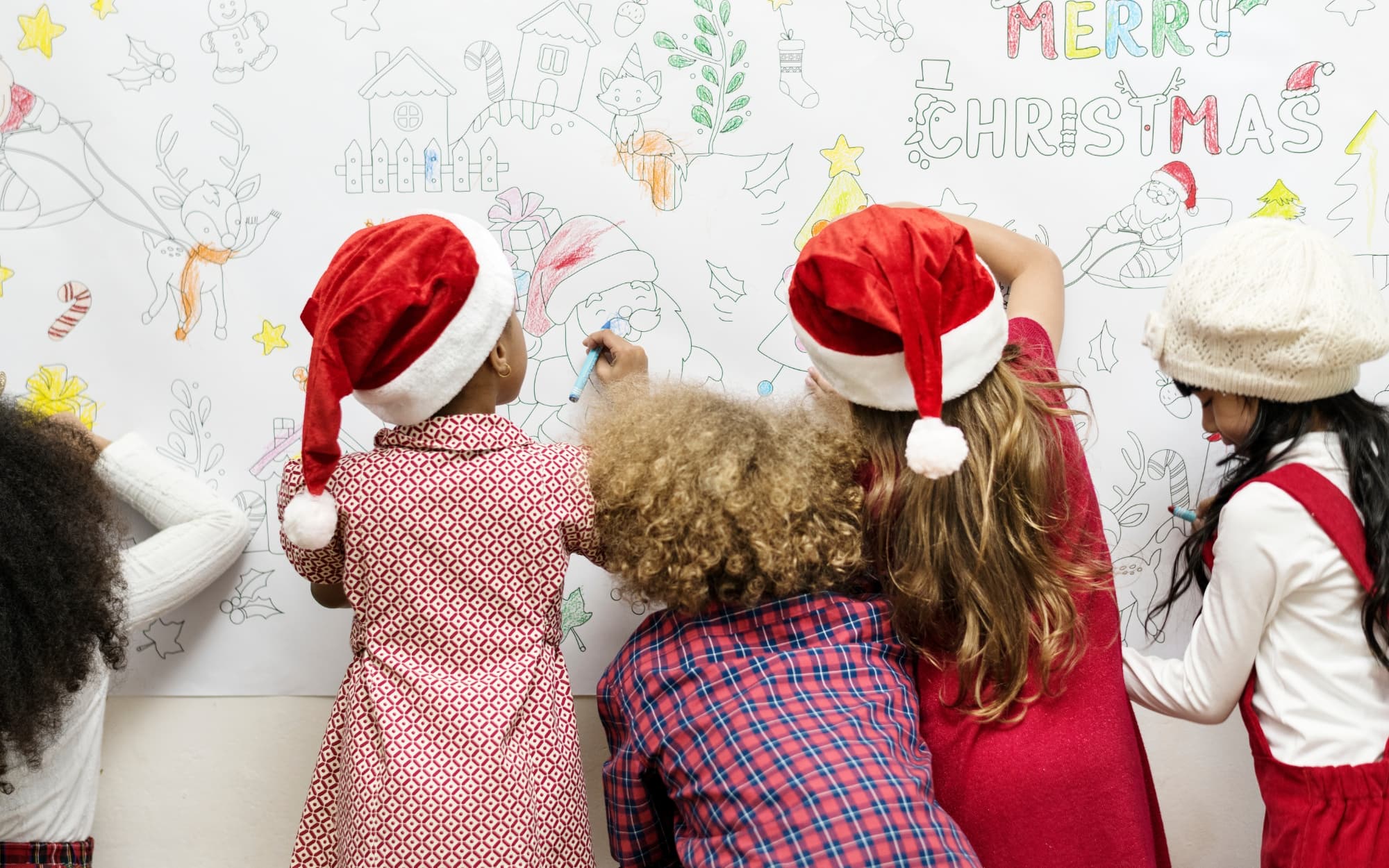 Children wearing Santa hats write Christmas messages on a whiteboard.