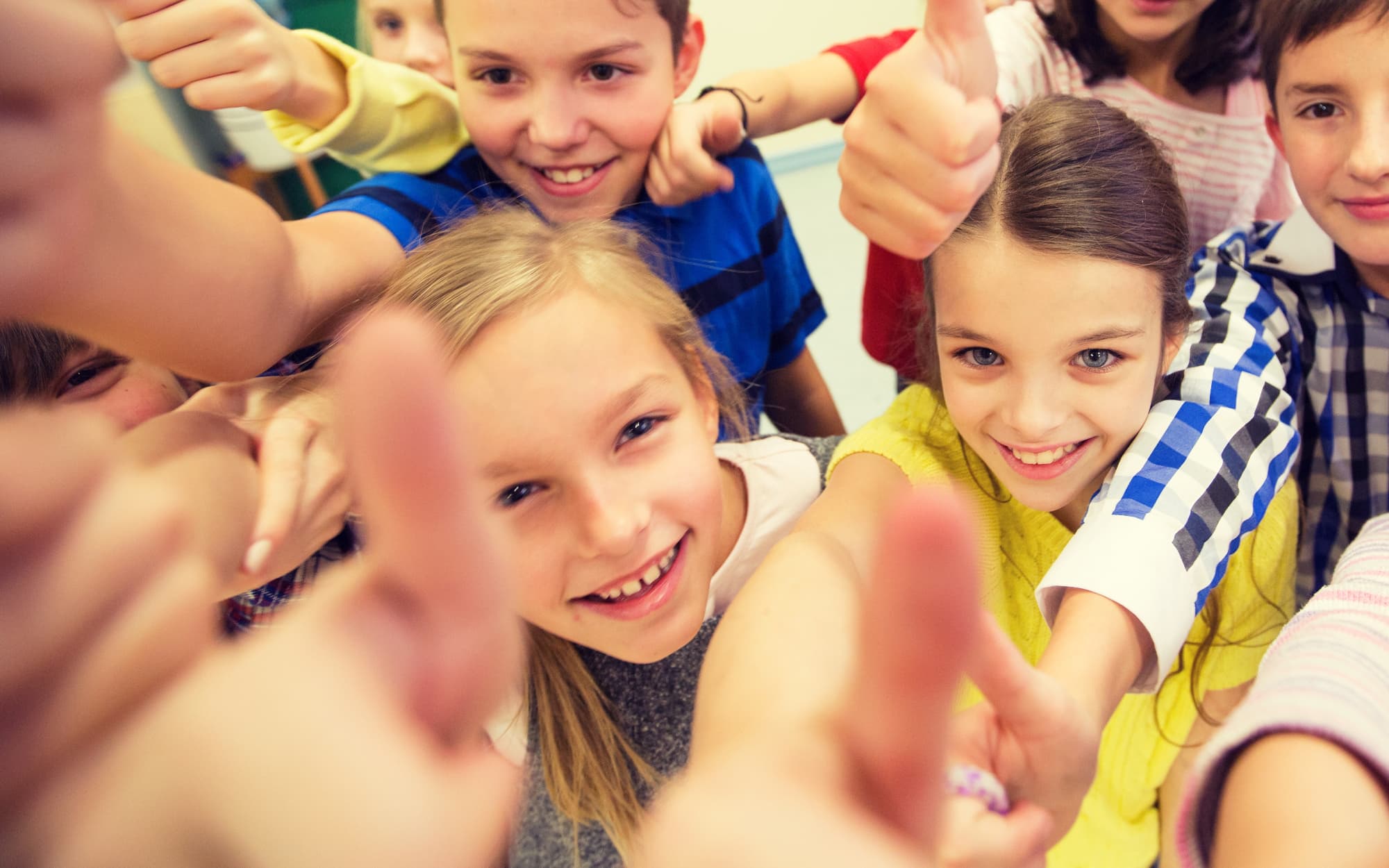 A classroom of children laugh and smile as they give thumbs-up to the camera.