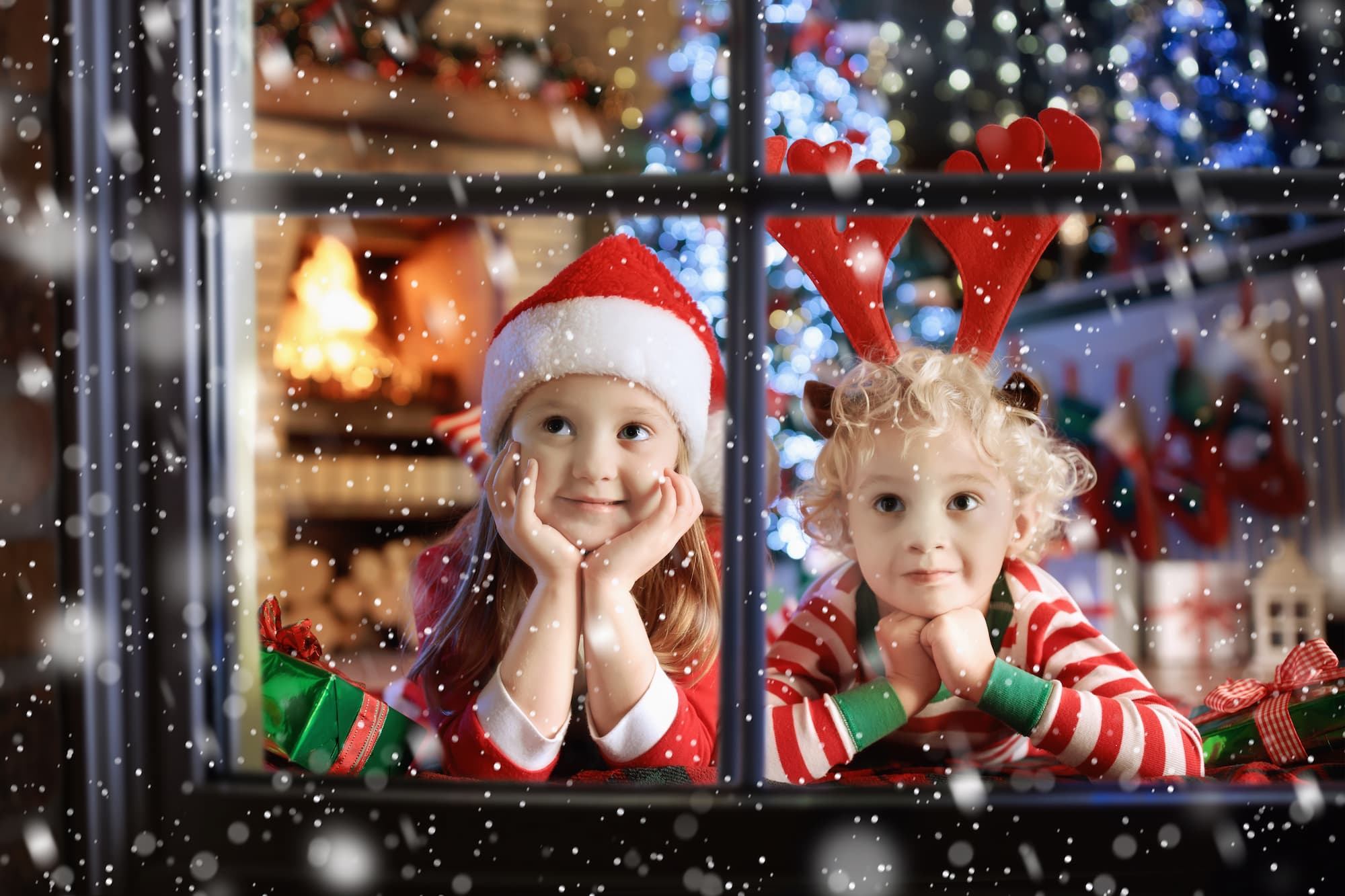 Two children wearing Santa hats smile while looking though a paned glass window on a snowy day.