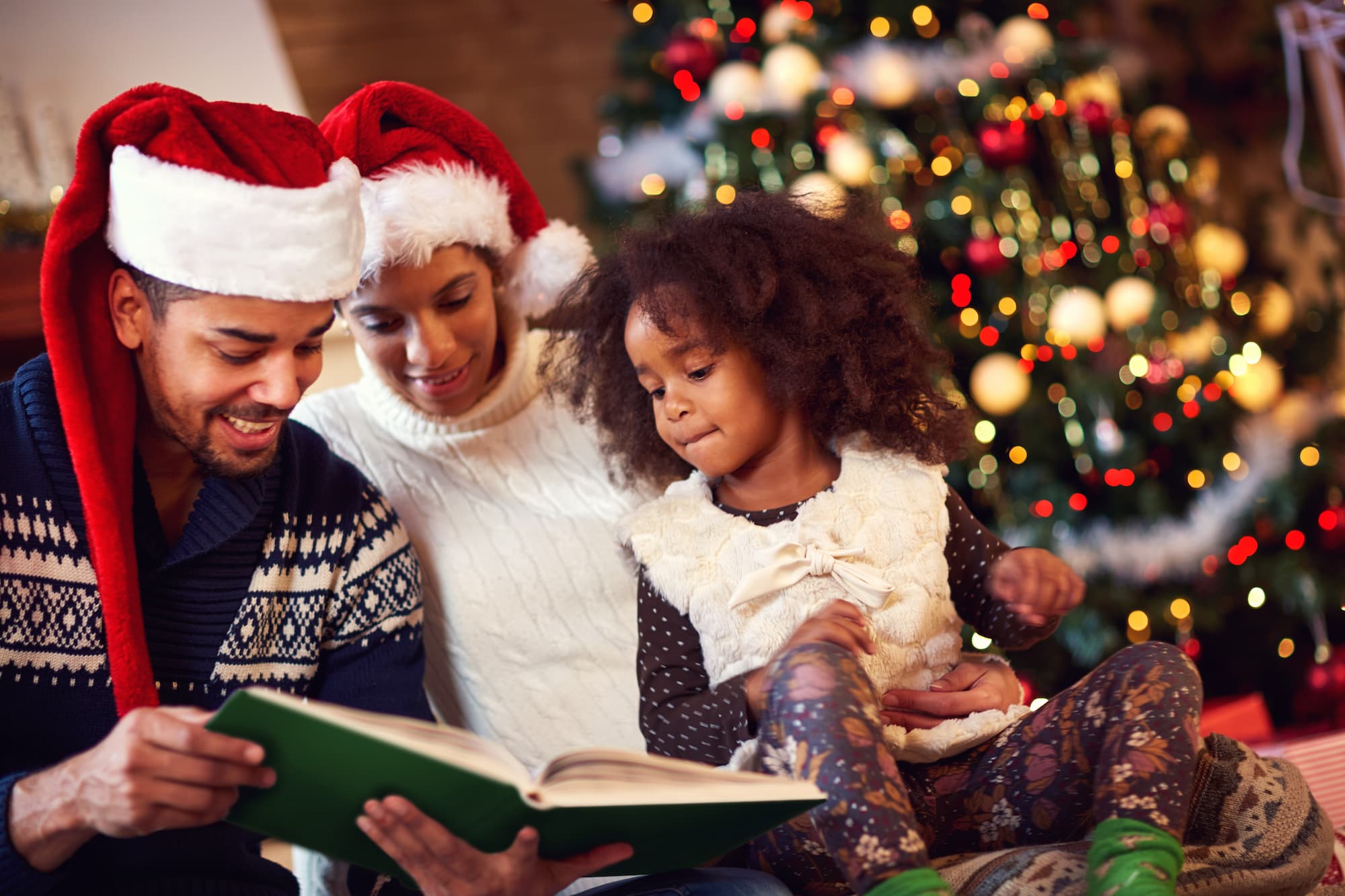 A family reads a Christmas book together while seated in front of an illuminated Christmas tree.