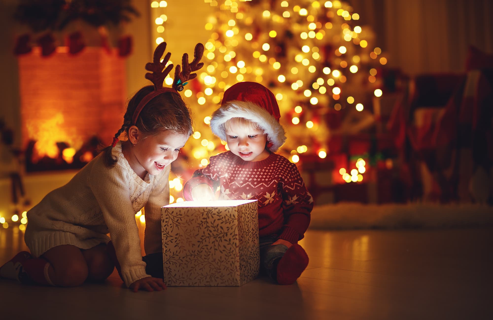 Two children open a Christmas present in front of an illuminated Christmas tree.