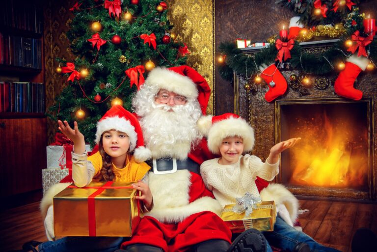 Tow children sit on Santa's lap, smiling and holding a present in front of a Christmas tree.