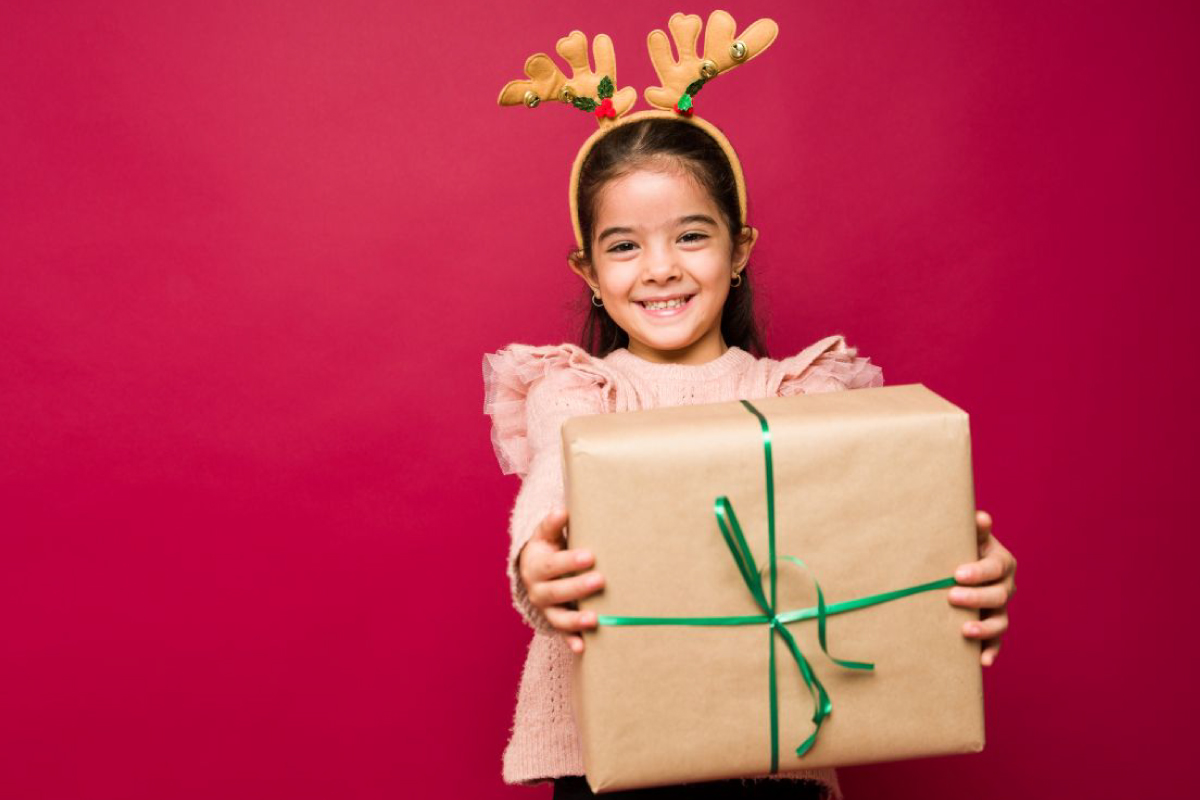 A cheerful child wearing festive antler headbands presents a beautifully wrapped holiday gift with a vibrant green ribbon, set against a bright red backdrop—perfectly capturing the joy and spirit of the season! Bring smiles like this to your school or organization’s fundraiser with our exclusive holiday catalog: filled with delightful gifts and seasonal treasures that families will love, all while supporting your fundraising goals.